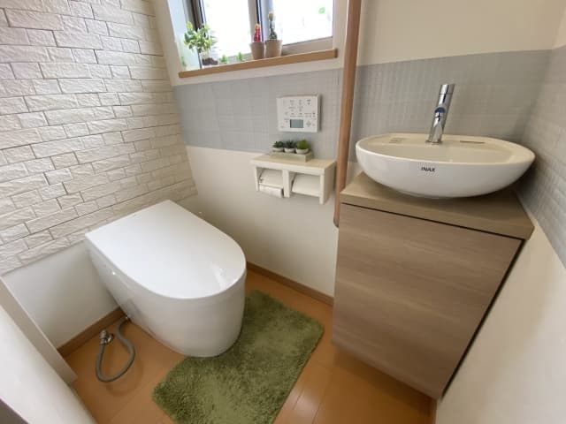 白いレンガ調の壁と明るい木目の手洗い収納を組み合わせた、清潔感あふれるナチュラルなトイレの内装事例