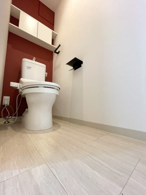 濃い赤色のアクセントクロスと、明るい石目調の床材を組み合わせたモダンなトイレの内装。白い便器と収納棚、黒いアクセサリー。