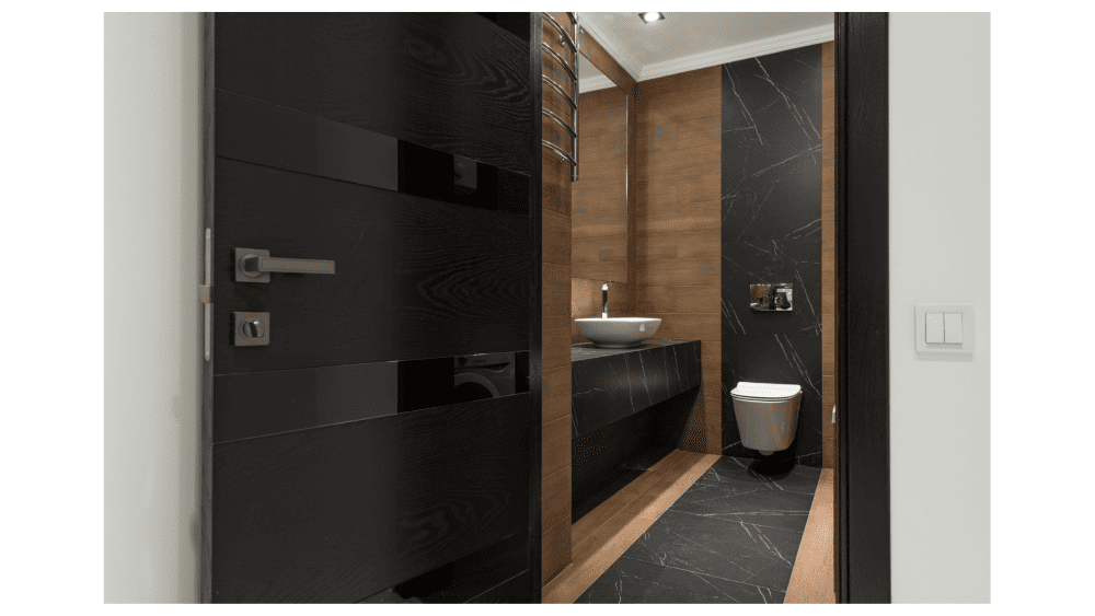 モダンなデザインのトイレ。手前に黒い扉があり、奥には黒の大理石調の床と木目の壁、白い洗面ボウルが見える清潔な空間。