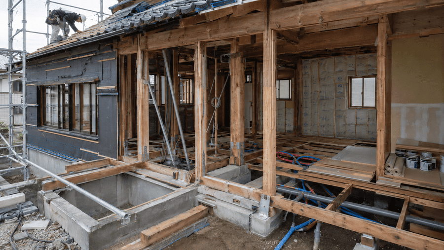 築古戸建てのリフォーム必要範囲がわかるスケルトン現場:基礎・柱・梁の構造体と床下配管、壁の断熱材が露出し、屋根では職人が補修作業を行っている様子