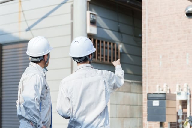 リフォームか建て替えかの判断に必要な住宅診断:ヘルメットをかぶった2人の専門業者が戸建て住宅の外壁を指差しながら確認している様子
